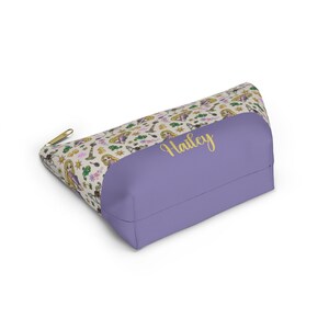 May include: A purple and cream-colored zippered pouch with a repeating pattern of a cartoon princess with long blonde hair. The pouch has a gold zipper and the name "Hailey" is written in gold on the front.