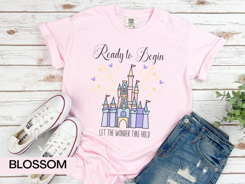 May include: Pink t-shirt with a black graphic of Cinderella's castle with fireworks and the text "Ready to Begin" and "Let the Wonder Take Hold".