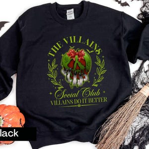 May include: Black sweatshirt with a green and red graphic of a skull with a bow, surrounded by leaves. Text reads "THE VILLAINS Social Club" and "VILLAINS DO IT BETTER". Halloween-themed apparel.