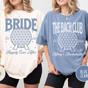 May include: Two t-shirts, one ivory and one blue jean, with a white graphic of a space mountain-like structure and text. The ivory shirt says "Bride" and "Happily Ever After". The blue jean shirt says "The Bach Club" and "Tiffany's Bachelorette".