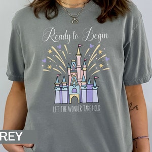 K&ouml;nnte beinhalten: Graues T-Shirt mit einem Motiv von Cinderellas Schloss und Feuerwerk. Der Text "Ready to Begin" und "Let the Wonder Take Hold" ist auf dem Shirt aufgedruckt.
