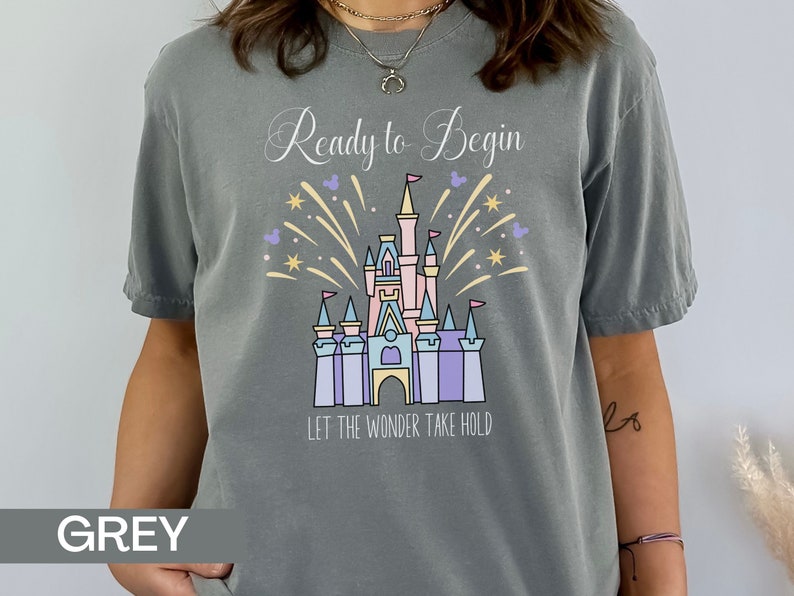 K&ouml;nnte beinhalten: Graues T-Shirt mit einem Motiv von Cinderellas Schloss und Feuerwerk. Der Text "Ready to Begin" und "Let the Wonder Take Hold" ist auf dem Shirt aufgedruckt.