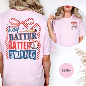 May include: Light pink t-shirt with a baseball-themed graphic. The back features the text "hey BATTER BATTER SWING!" in red, white, and blue, with a baseball and bat design. The front has a smaller baseball bow design with the name "MASON".