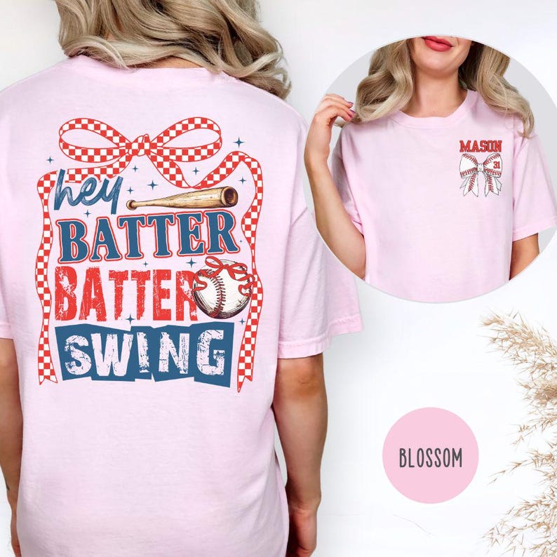 May include: Light pink t-shirt with a baseball-themed graphic. The back features the text "hey BATTER BATTER SWING!" in red, white, and blue, with a baseball and bat design. The front has a smaller baseball bow design with the name "MASON".