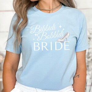 May include: Light blue t-shirt with the text "Bibbidi Bobbidi Bride" in white cursive font and a white glass slipper graphic.