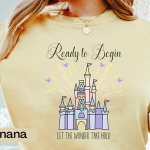 May include: A yellow t-shirt with a graphic of Cinderella's castle and fireworks. The text "Ready to Begin" and "Let the Wonder Take Hold" is printed on the shirt.