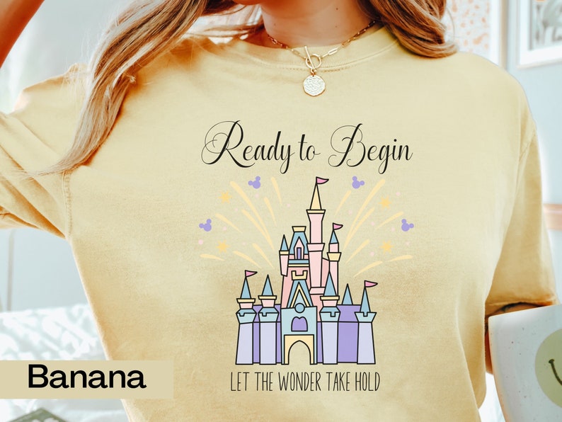 May include: A yellow t-shirt with a graphic of Cinderella's castle and fireworks. The text "Ready to Begin" and "Let the Wonder Take Hold" is printed on the shirt.