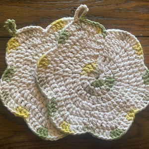 Puede incluir: Dos agarraderas de olla de ganchillo con forma de flores. Las agarraderas son blancas con detalles verdes y amarillos.