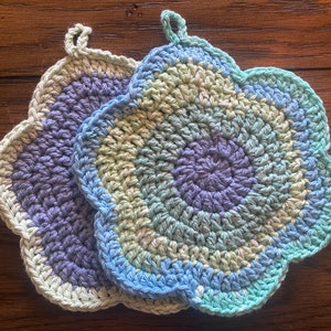 May include: Two crocheted flower-shaped pot holders in shades of blue, green, and purple. The pot holders are made with a textured stitch and have a scalloped edge.
