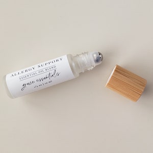 May include: A clear glass roller bottle with a silver roller ball. The bottle is labeled "Allergy Support Essential Oil Blend grace essentials 1/3 oz | 10 ml". The bottle is sitting on a white surface with a wooden cap next to it.