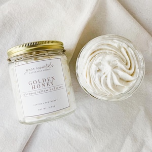May include: Two glass jars of whipped body butter on a white cloth. One jar is closed with a gold lid and a label that reads "GOLDEN HONEY whipped tallow bodycare." The other jar is open, revealing a swirl of creamy, white body butter. Net wt. 3.5oz.