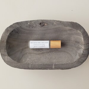 May include: A small, white bottle with a wooden roller ball top. The bottle is labeled "HEADACHE RELIEF ESSENTIAL OIL BLEND grace essentials". The bottle is sitting in a gray wooden bowl.