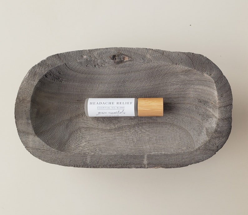 May include: A small, white bottle with a wooden roller ball top. The bottle is labeled "HEADACHE RELIEF ESSENTIAL OIL BLEND grace essentials". The bottle is sitting in a gray wooden bowl.