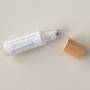 May include: A clear glass roller bottle with a silver roller ball. The bottle is labeled "STRESS BE GONE ESSENTIAL OIL BLEND grace essentials 1/3 oz | 10 ml". The bottle is sitting on a white surface with a wooden cap next to it.