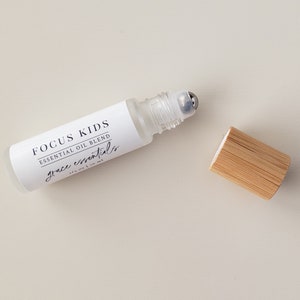 May include: A clear glass roller bottle with a white label that reads "FOCUS KIDS ESSENTIAL OIL BLEND grace essentials 1/3 oz | 10 ml". The bottle has a silver roller ball and a bamboo lid.