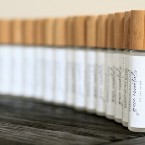 May include: A row of small glass bottles with bamboo lids and white labels. The label on the front bottle reads "grace essentials" and lists the ingredients: "peace and calming oil, lemon oil, frankincense oil, organic jojoba carrier oil". The label also states "1/3 oz | 10 ml".