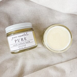 May include: A small glass jar with a white lid filled with a yellow balm. The jar has a label that reads "grace essentials PURE solid tallow balm unscented net wt. 2oz".