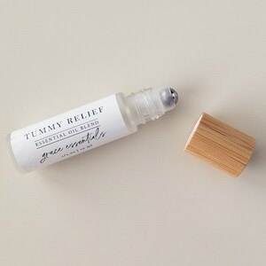 May include: A clear glass roller bottle with a white label that reads "Tummy Relief Essential Oil Blend Grace essentials 1/3 oz | 10 ml". The bottle has a silver roller ball and is sitting on a white surface. A wooden cap is next to the bottle.