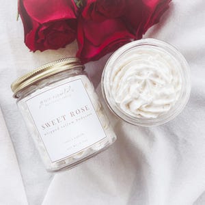 May include: A jar of whipped tallow bodycare labeled "Sweet Rose" with rose and vanilla scent. The jar is open, revealing the creamy product. Two red roses are in the background.
