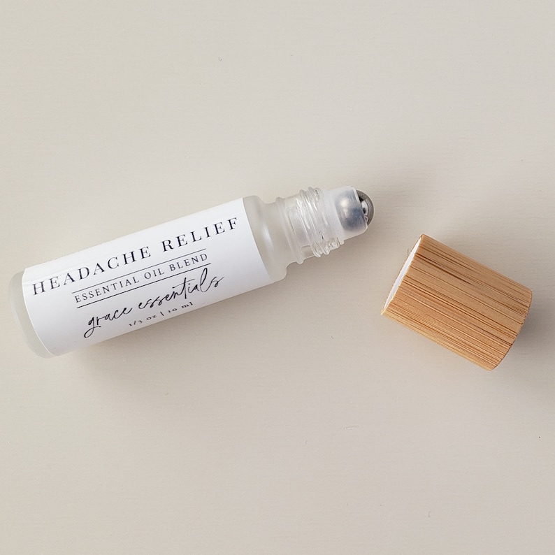 May include: A clear glass roller bottle with a silver roller ball. The bottle is labeled "HEADACHE RELIEF ESSENTIAL OIL BLEND grace essentials 1/3 oz | 10 ml". The bottle is sitting on a light beige surface with a wooden cap next to it.