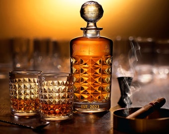 May include: A clear glass decanter with a diamond-shaped pattern and gold lettering that reads "John Roberts Est. 2023". The decanter is filled with amber liquid and has a round stopper. Two clear glass tumblers with a diamond-shaped pattern are in front of the decanter. A cigar and a small glass are also in the image.