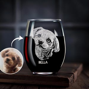 May include: Stemless wine glass with a detailed etched portrait of a dog, with the name "BELLA" below the image. The glass is filled with red liquid. A smaller circular image of the dog is next to the glass. The glass sits on a wooden surface.