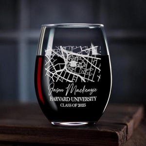 May include: A stemless wine glass filled with red liquid. The glass is etched with a map design and the text "Jason Mackenzie, Harvard University, Class of 2025". The glass sits on a dark wooden surface.