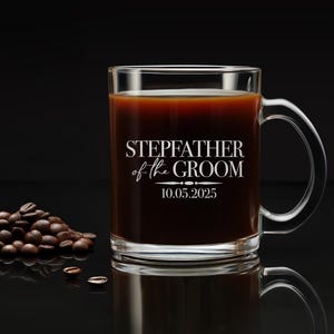 以下が含まれることがあります： 黒い取っ手のある透明なガラス製のコーヒーカップ。カップには「Stepfather of the Groom」という文字と「10.05.2025」という日付が印刷されています。カップはコーヒーで満たされ、黒い表面の上に置かれています。