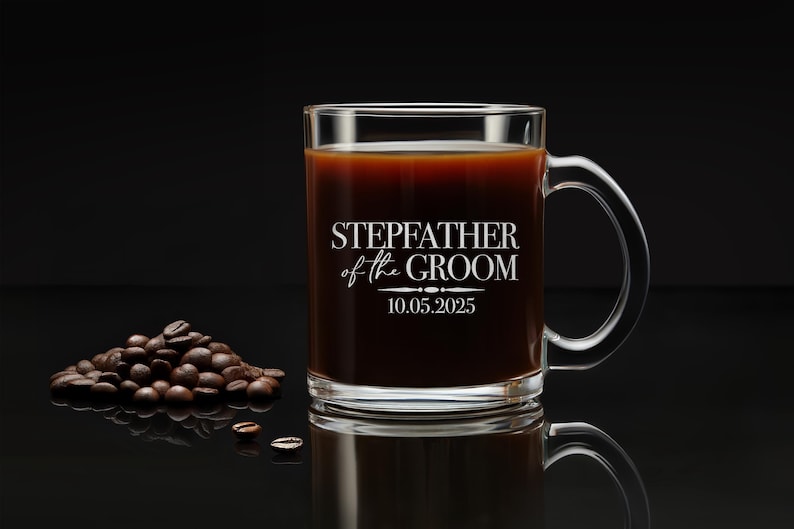 以下が含まれることがあります： 黒い取っ手のある透明なガラス製のコーヒーカップ。カップには「Stepfather of the Groom」という文字と「10.05.2025」という日付が印刷されています。カップはコーヒーで満たされ、黒い表面の上に置かれています。