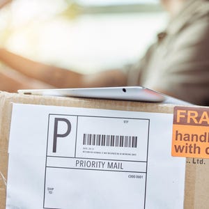 May include: A cardboard box with a white label marked "PRIORITY MAIL" and a barcode. An orange sticker reads "FRAGILE handle with care." A silver tablet rests on top of the box. The box is in a vehicle.