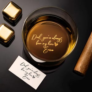 May include: A rocks glass filled with amber liquid, featuring the engraved text "Dad, you've always been my hero Erica." Two gold-colored square ice cubes and a cigar are also present. A small card with the same text is next to the glass.