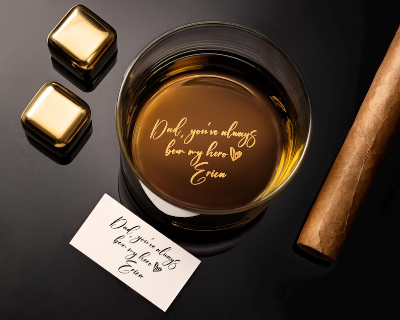May include: A rocks glass filled with amber liquid, featuring the engraved text "Dad, you've always been my hero Erica." Two gold-colored square ice cubes and a cigar are also present. A small card with the same text is next to the glass.