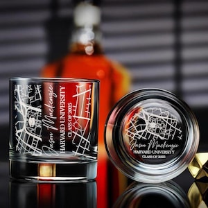 May include: Two clear, personalized whiskey glasses with a map design and the text "Jason Mackenzie, Harvard University, Class of 2025." One glass is upright, the other is on its side. A bottle is blurred in the background.