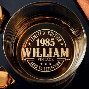 May include: A whiskey glass with a personalized design, featuring the name "WILLIAM" and the year "1985" in gold lettering. The glass is filled with amber liquid, accompanied by gold ice cubes and a cigar.