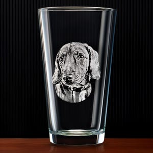 May include: A clear pint glass with a detailed etched image of a dog's face. The dog has floppy ears and a collar. The glass is on a wooden surface with a black background.
