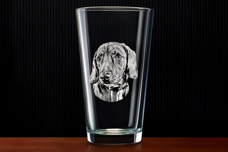 May include: A clear pint glass with a detailed etched image of a dog's face. The dog has floppy ears and a collar. The glass is on a wooden surface with a black background.