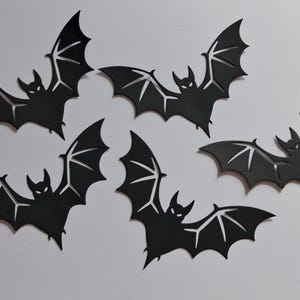 May include: A set of five black bat-shaped cutouts featuring pointed ears and detailed wing structures with cut-out sections. Each bat is depicted in a flying position with wings spread wide, showcasing a flat, matte black finish and sharp edges.