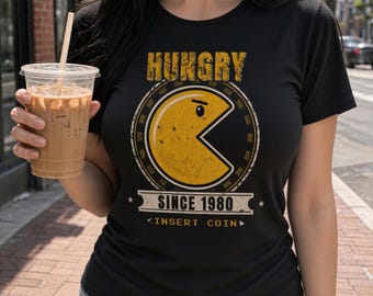 May include: A black short-sleeve t-shirt featuring a yellow circular character graphic with a wedge-shaped mouth. The design includes the text HUNGRY, SINCE 1980, and INSERT COIN in a distressed font style, centered on the chest area.