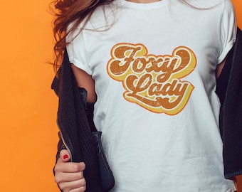 May include: White t-shirt with a retro-style graphic that says "Foxy Lady" in orange and yellow lettering.