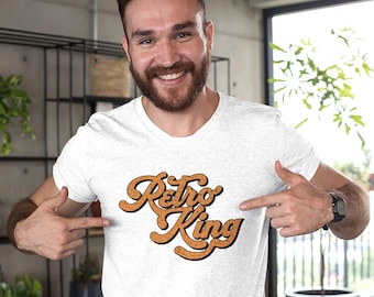 May include: A light gray heather crew neck t-shirt featuring the text Retro King printed in a bold, orange, cursive font with a dark outline. The lettering has a textured, speckled appearance across the chest of the garment.