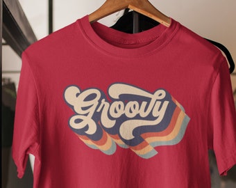 May include: A red t-shirt with the word "Groovy" in a retro font. The word is in white with a blue shadow and rainbow stripes. The shirt is hanging on a wooden hanger.