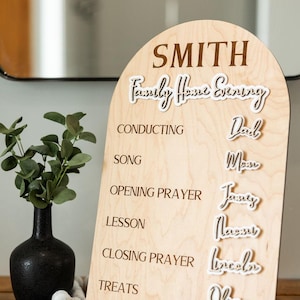 May include: Wooden family home evening schedule board with the family name "Smith" at the top. The board lists activities like conducting, song, and lesson. Names of family members are listed next to each activity. A black vase with greenery is in the background.