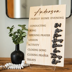 May include: A wooden family home evening sign with the family name "Anderson" at the top. The sign lists activities like "Conducting" and "Song", with family member names in black script. A black vase with greenery sits to the left.