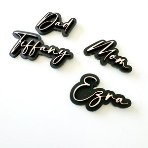 以下が含まれることがあります： 金色の文字で書かれた黒い木製のネームタグが4つ。名前は「お父さん」、「ティファニー」、「お母さん」、「エズラ」です。