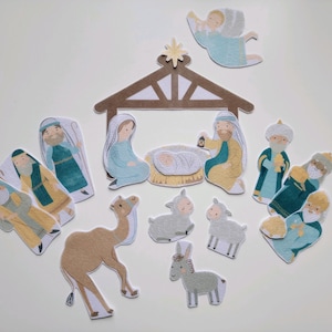Puede incluir: Un conjunto de recortes de papel que representan una escena de la Natividad. El conjunto incluye figuras de María, José, el niño Jesús, ángeles, reyes magos y animales. La paleta de colores presenta azules, amarillos y marrones suaves.