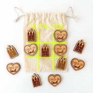 May include: A wooden tic-tac-toe game with a beige canvas bag. The game pieces are wooden hearts with the words "Child of God" and wooden temple silhouettes.