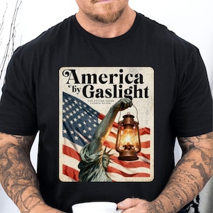 May include: Black t-shirt featuring a graphic with the text "America by Gaslight." The design includes the Statue of Liberty holding a lantern against a backdrop of the American flag. The text "The Future Never Looked So Dim" is also included.