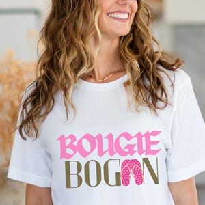 May include: White t-shirt with the text "Bougie Bogan" in pink and brown lettering. The text is in a stylized font. The shirt also features a pair of pink flip flops with white polka dots.