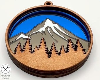 Peut inclure: Une décoration ronde en bois avec un design en couches représentant une chaîne de montagnes avec des sommets enneigés et un ciel bleu. La chaîne de montagnes est entourée d'une bordure bleu foncé.