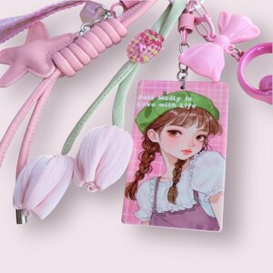 May include: A collection of pink accessories, including a star-shaped charm, a bow, and a keychain with a cartoon girl illustration. The illustration features a girl with brown braids and a green beret. The text on the keychain reads "Fall Madly in Love with Life."